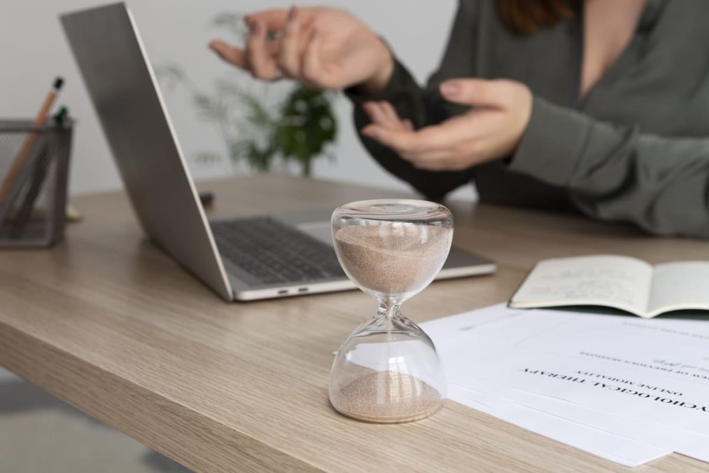Mesa de trabalho com uma ampulheta em primeiro plano, simbolizando a gestão do lead time em processos, enquanto uma profissional realiza uma sessão ou reunião ao fundo.