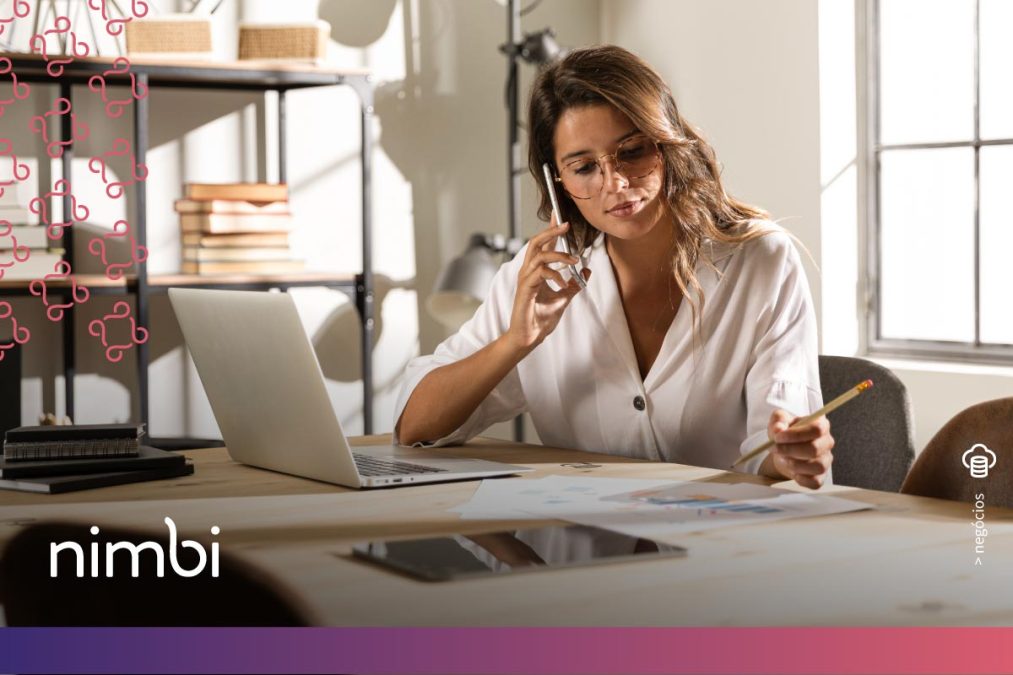 Uma mulher usando óculos está sentada em uma mesa de trabalho, falando ao telefone enquanto segura um lápis e olha para alguns documentos. Ela tem um laptop aberto à sua frente, demonstrando as habilidades de um comprador em um ambiente de trabalho focado e produtivo. No canto inferior esquerdo da imagem, o logotipo da Nimbi é exibido.