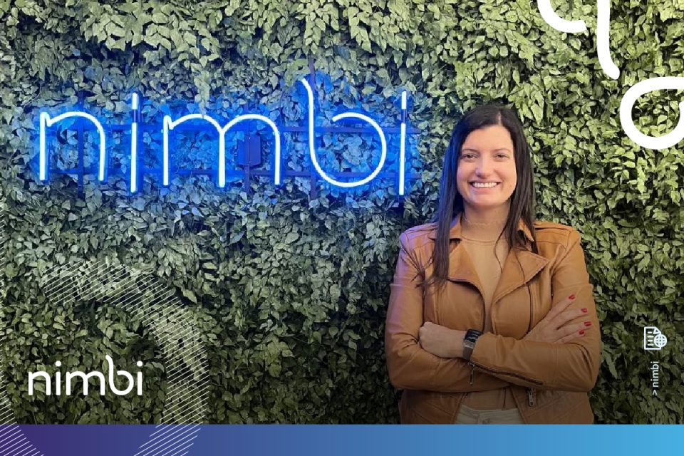 Mulher branca vestindo jaqueta marro, CEO da Nimbi Carolina Cabral. Ela está em fundo verde com luminária escrito "nimbi"