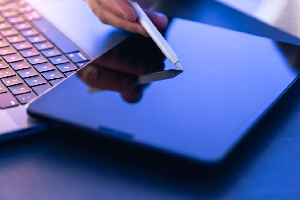 Close-up de uma pessoa utilizando uma caneta stylus para realizar uma assinatura eletrônica em um tablet, com um laptop ao lado, em um ambiente de trabalho digital.