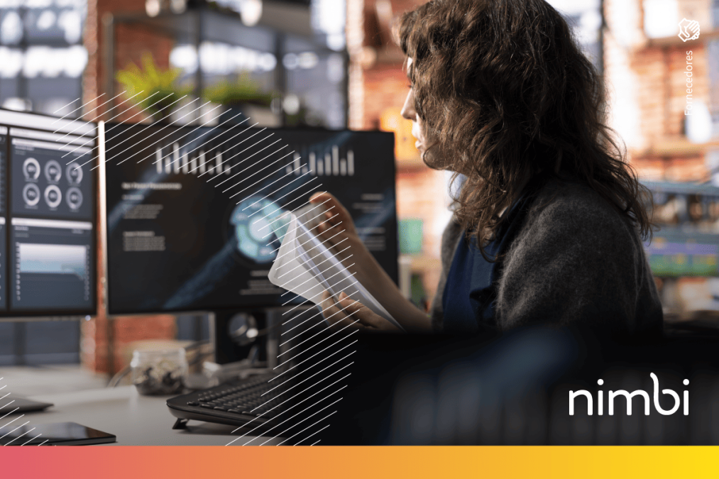 Mulher em escritório analisa documentos físicos e gráficos em múltiplos monitores, visualizando o fluxo de dados transacionais.