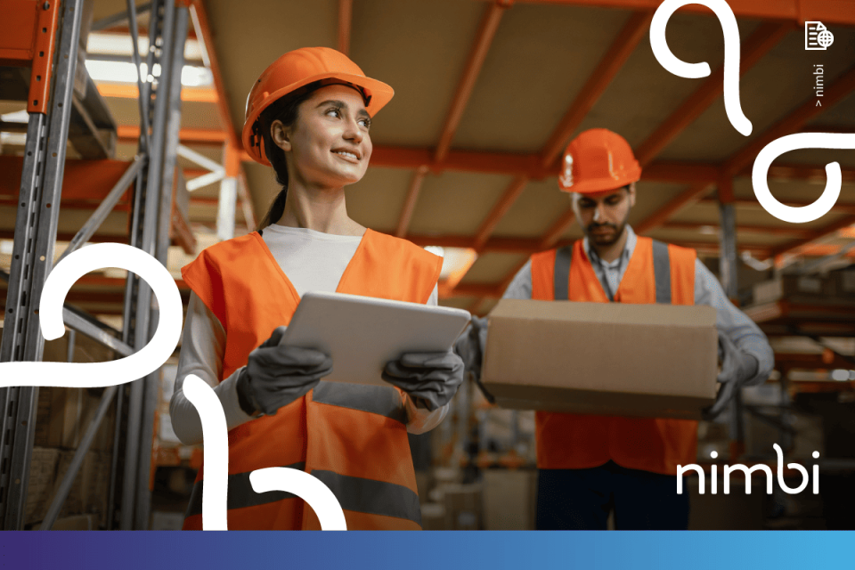 Trabalhadora com capacete e colete laranja segura tablet em armazém com colega ao fundo, ilustrando a gestão de segurança e normas de QHSE.